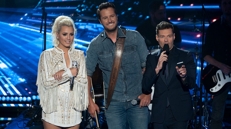 Gabby Barrett in a white dress stands holding a microphone on the "American Idol" stage with Luke Bryan in all denim and Ryan Seacrest