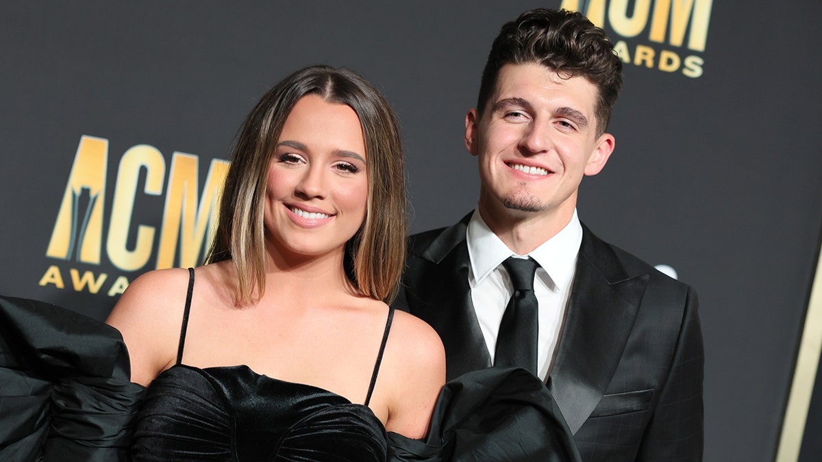 Gabby Barrett in a black dress smiles next to husband Cade Foehner in a black suit and tie at the Academy Of Country Music Awards