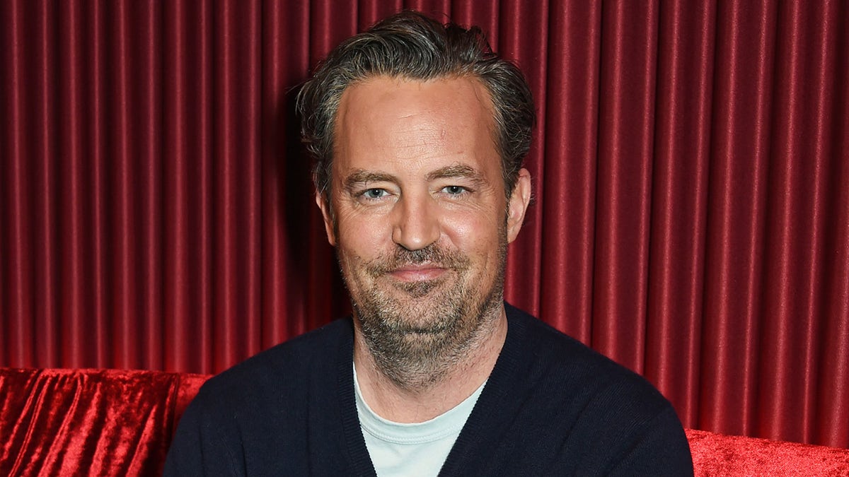 Matthew Perry in a black sweater soft smiles in front of red curtain