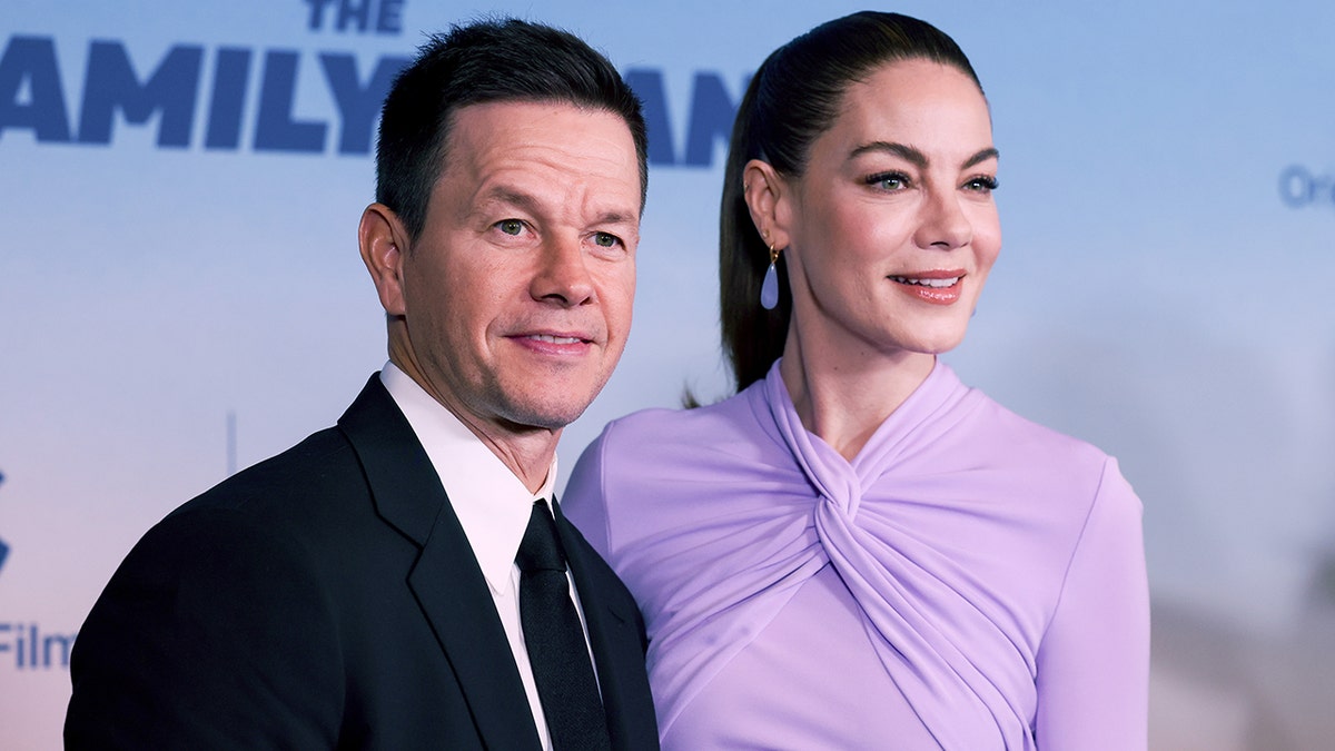 Mark Wahlberg in a dark suit and tie stands next to Michelle Monaghan in a lilac dress